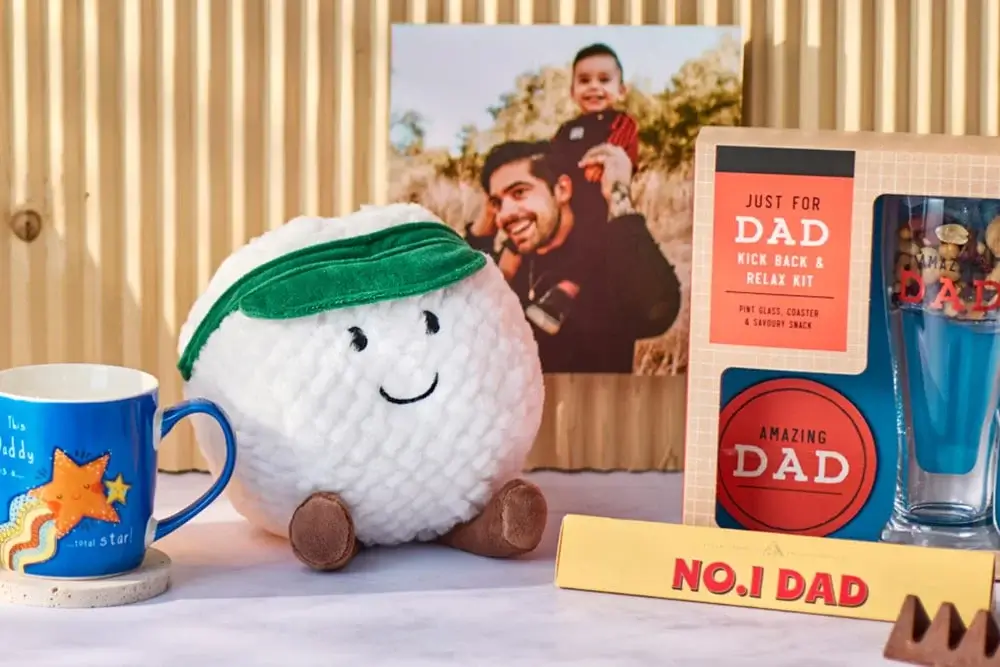 'The image shows a collection of Father's Day gifts arranged on a table. There is a blue mug with the text 'You are my star' and an illustration of a star, a plush toy resembling a golf ball with a green visor and brown feet, and a boxed gift set labeled 'JUST FOR DAD KICK BACK & RELAX KIT' which includes pint glass coasters and leather socks. Additionally, there is another boxed item labeled 'AMAZING DAD' containing what appears to be shot glasses. A wooden block with the text 'NO.1 DAD' is also present. In the background, there is a framed photo of an adult holding a child.' 'The image shows a collection of Father's Day gifts arranged on a table. There is a blue mug with the text 'You are my star' and an illustration of a star, a plush toy resembling a golf ball with a green visor and brown feet, and a boxed gift set labeled 'JUST FOR DAD KICK BACK & RELAX KIT' which includes pint glass coasters and leather socks. Additionally, there is another boxed item labeled 'AMAZING DAD' containing what appears to be shot glasses. A wooden block with the text 'NO.1 DAD' is also present. In the background, there is a framed photo of an adult holding a child.'