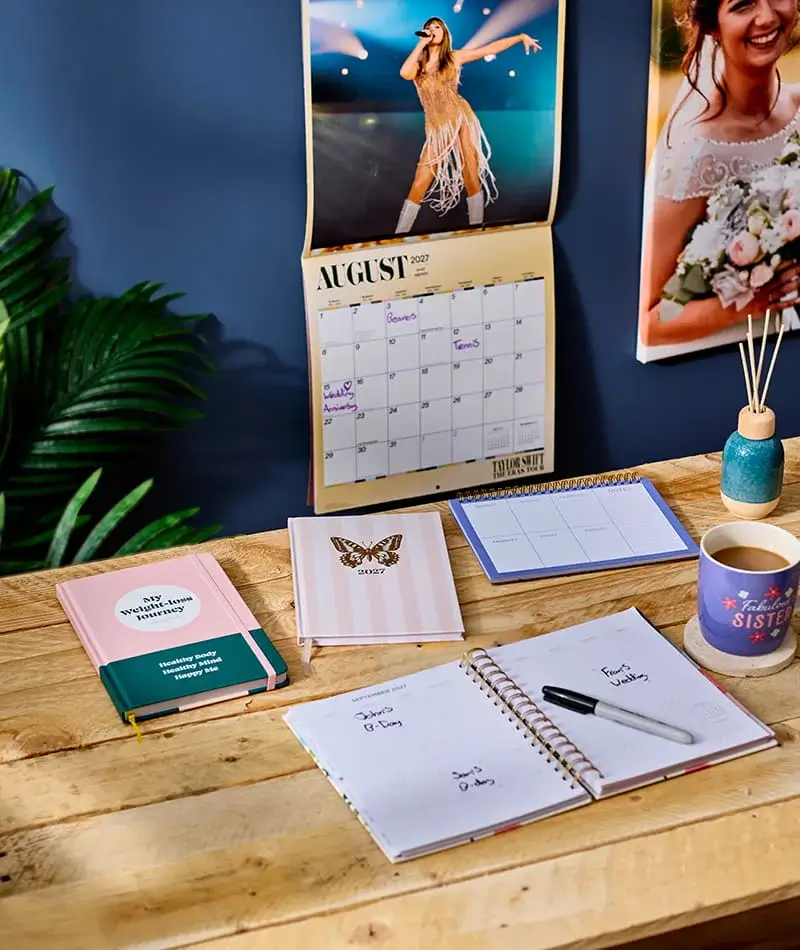 Desk stationery collection featuring a personalised wall calendar, notebooks, weekly planner, diffuser, and mug arranged on a wooden desk.