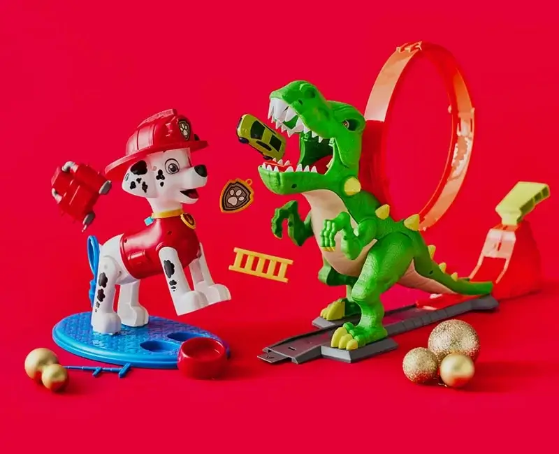 Toy Dalmatian firefighter and green dinosaur on a red background with a loop track, flying toy cars, firefighting accessories, and gold Christmas ornaments. Toy Dalmatian firefighter and green dinosaur on a red background with a loop track, flying toy cars, firefighting accessories, and gold Christmas ornaments.