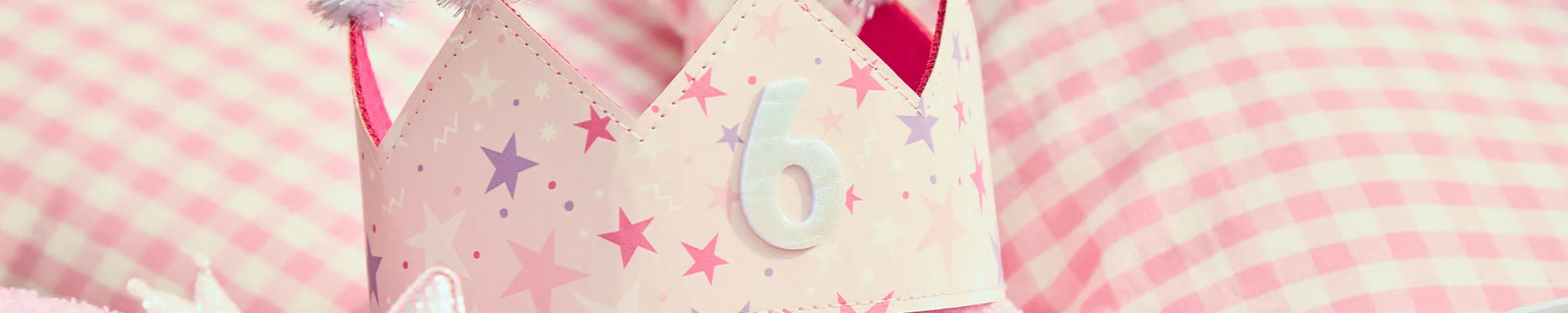 A close-up of a pastel pink princess crown decorated with star patterns and topped with fluffy pom-poms, featuring a white number 6 on the front. The crown is displayed against a pink gingham cushion background. A close-up of a pastel pink princess crown decorated with star patterns and topped with fluffy pom-poms, featuring a white number 6 on the front. The crown is displayed against a pink gingham cushion background.