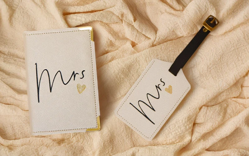 A white passport holder and matching luggage tag set, both printed with ‘Mrs’ in black script and a small gold heart, displayed on soft beige fabric. A white passport holder and matching luggage tag set, both printed with ‘Mrs’ in black script and a small gold heart, displayed on soft beige fabric.