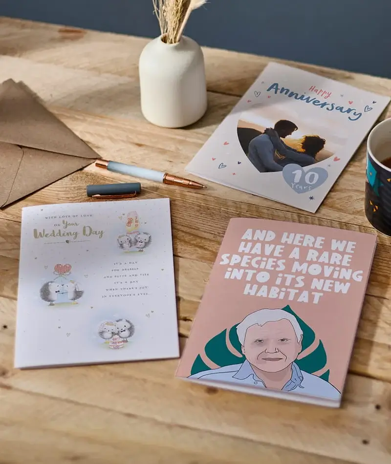 Selection of greeting cards including anniversary, wedding day, and humorous designs arranged on a wooden table with stationery and a mug. Selection of greeting cards including anniversary, wedding day, and humorous designs arranged on a wooden table with stationery and a mug.