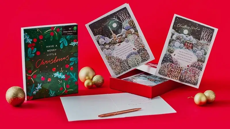 Set of Christmas cards on a bright red background, including a box labeled Have a Merry Little Christmas with festive foliage and berries, two propped-up cards showing snowy winter scenes with a church and a house, gold ornaments, and an open card with an envelope and pen.