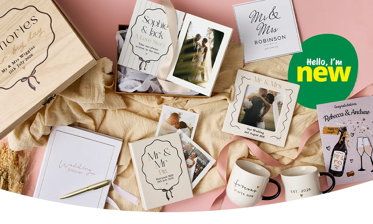 A flat lay of coordinated wedding gifts and keepsakes including a wooden memories box, personalised cards, photo prints, mugs, a notebook, ribbons and decorative foliage arranged on a soft pink background.