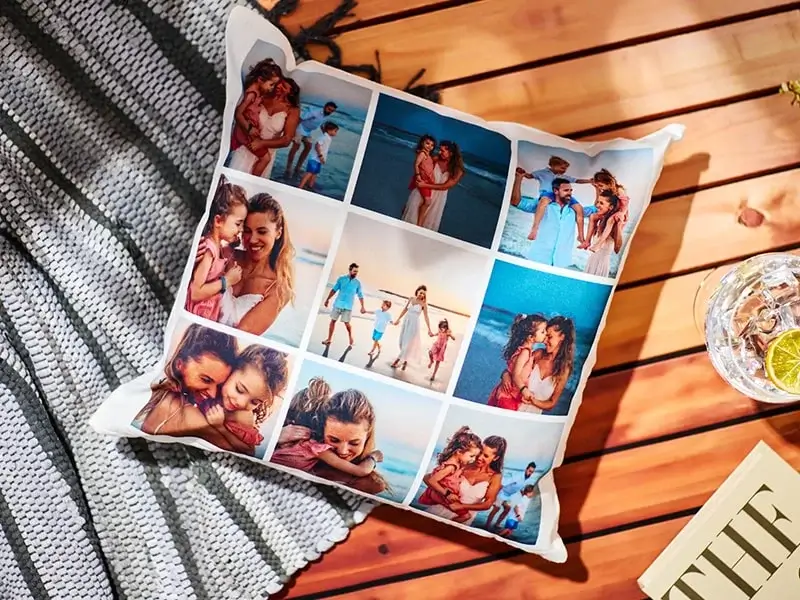 A decorative pillow featuring a collage of nine photos showing various moments of a family at the beach. The images include scenes of a couple embracing, children playing, and family members walking along the shore. The pillow is placed on a wooden surface next to a striped blanket and a glass with lime slices.