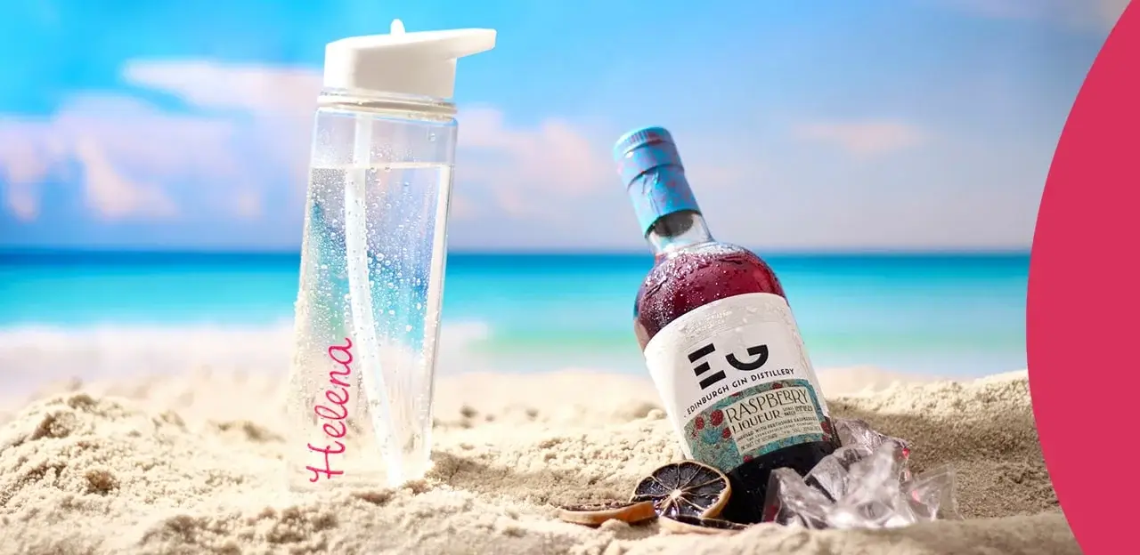 A clear water bottle with the name Helena written in pink is placed on sandy beach next to a bottle of Edinburgh Gin Distillery Raspberry Liqueur. The background shows a serene ocean view with blue skies and soft clouds. There are also some ice cubes and dried citrus slices near the gin bottle. A clear water bottle with the name Helena written in pink is placed on sandy beach next to a bottle of Edinburgh Gin Distillery Raspberry Liqueur. The background shows a serene ocean view with blue skies and soft clouds. There are also some ice cubes and dried citrus slices near the gin bottle.