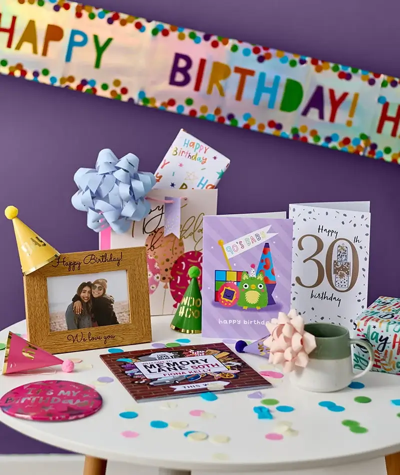 Birthday celebration display featuring greeting cards, a personalised photo frame, wrapped gifts, party hats and confetti, set against a purple backdrop with a birthday banner. Birthday celebration display featuring greeting cards, a personalised photo frame, wrapped gifts, party hats and confetti, set against a purple backdrop with a birthday banner.