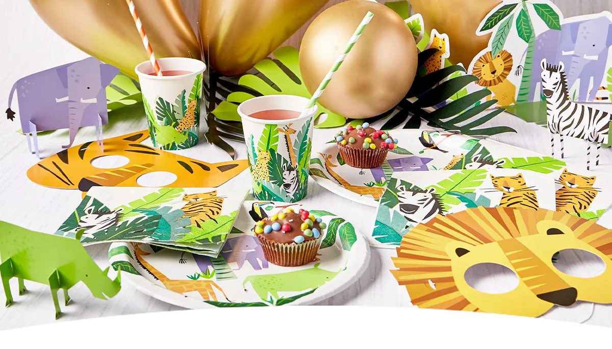 A close-up safari party table featuring animal-print tableware with zebra, tiger and elephant designs, surrounded by tropical leaf decorations, gold balloons and animal-shaped table centrepieces. Lion face masks, jungle-themed cups and decorated cupcakes complete the coordinated jungle celebration display.