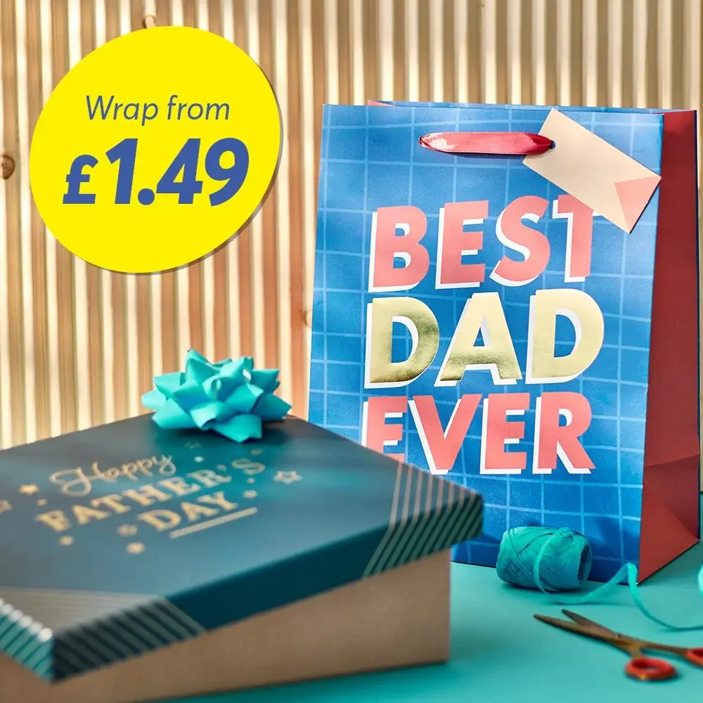 A gift bag with the text 'BEST DAD EVER' in bold letters and a price tag that reads 'Wrap from £1.49'. Next to the bag is a gift box with a blue ribbon and the text 'Happy Father's Day'. There is also a spool of blue ribbon on the table.