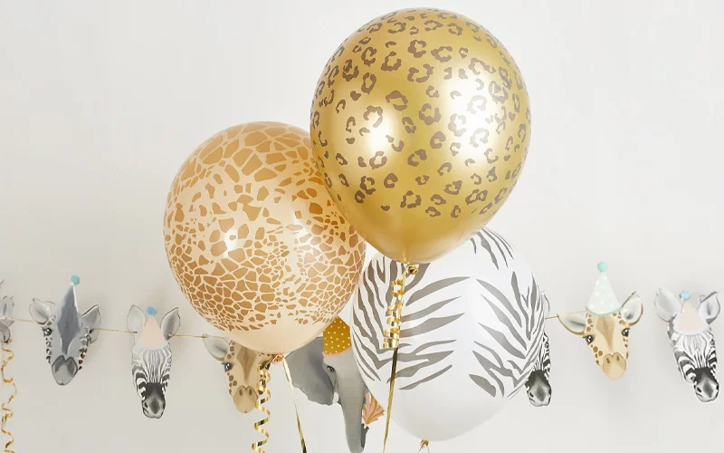 A cluster of safari-themed balloons featuring metallic gold, giraffe-print and zebra-print designs, displayed in front of animal-face bunting with zebra, giraffe, elephant and leopard illustrations wearing party hats.