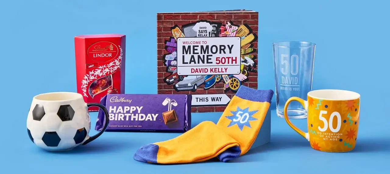 'The image shows a collection of 50th birthday-themed items arranged on a blue background. The items include a soccer ball mug, a box of Lindt Lindor milk chocolates, a Cadbury Happy Birthday chocolate bar, a pair of yellow and blue socks with the number 50 on them, an orange mug with the text '50 No intention of acting my age,' and a clear glass with the text '50 David beers to you!' There is also a book titled 'Welcome to Memory Lane 50th' by David Kelly.'