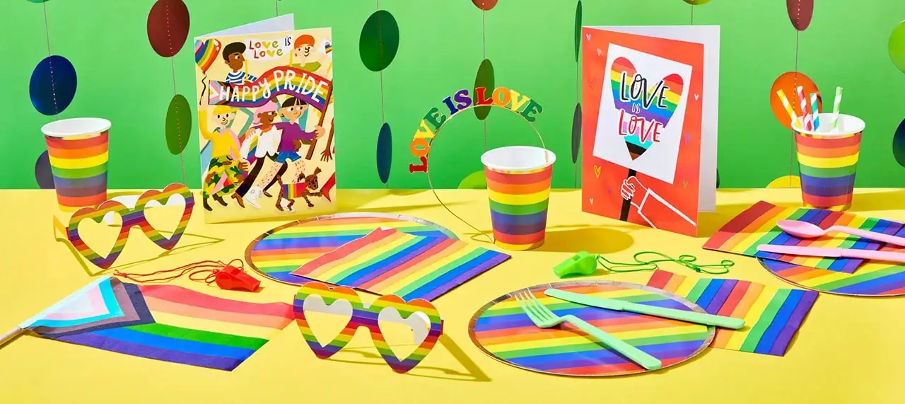 'The image shows a colorful Pride-themed party setup with rainbow decorations. There are two greeting cards, one with the text 'Love is Love' and another with 'Happy Pride'. The table is adorned with rainbow-striped plates, cups, napkins, and utensils. Heart-shaped glasses and a headband with the text 'Love is Love' are also present. The background features hanging circular decorations in various colors.'