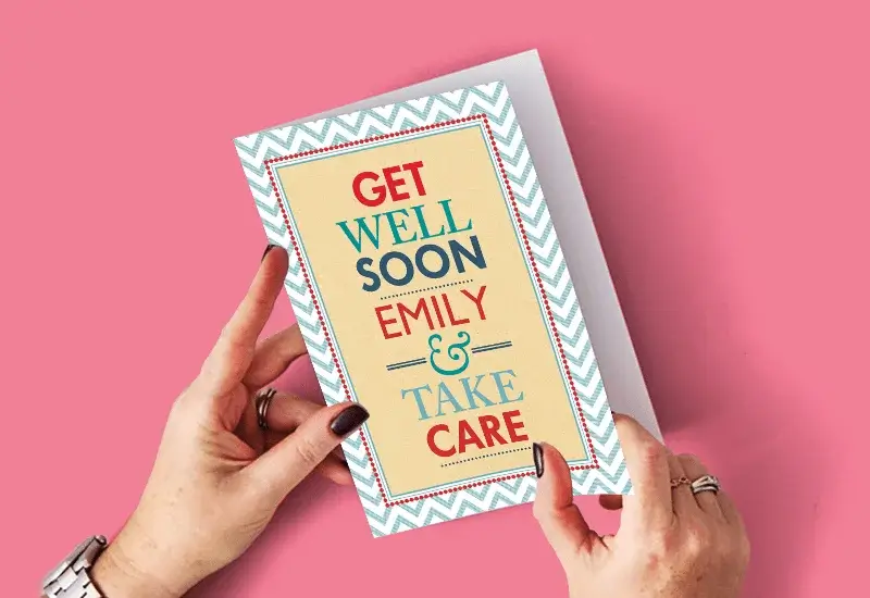 A person holding a get well soon card with a chevron border on a pink background and bold colorful text saying GET WELL SOON EMILY and TAKE CARE.