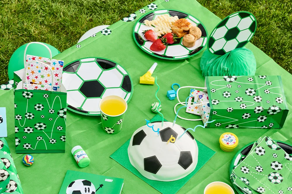 A football-themed birthday party setup on a green tablecloth, featuring a football-shaped cake, football-print plates, cups and napkins, party favour bags, gift bags and small party bag fillers such as bubbles, slime and toys. A football honeycomb decoration and football-patterned tableware complete the coordinated party display. A football-themed birthday party setup on a green tablecloth, featuring a football-shaped cake, football-print plates, cups and napkins, party favour bags, gift bags and small party bag fillers such as bubbles, slime and toys. A football honeycomb decoration and football-patterned tableware complete the coordinated party display.