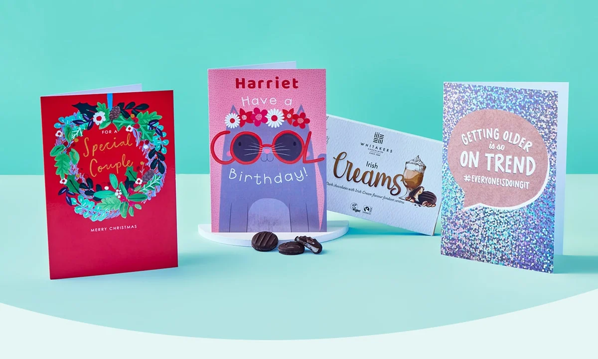 Four greeting cards and a box of chocolates arranged on a light blue surface with a teal background, including a red Christmas card with a wreath, a pink birthday card with sunglasses and flowers, a box of Whitakers Irish Creams, and a silver glittery card with a humorous message about getting older.