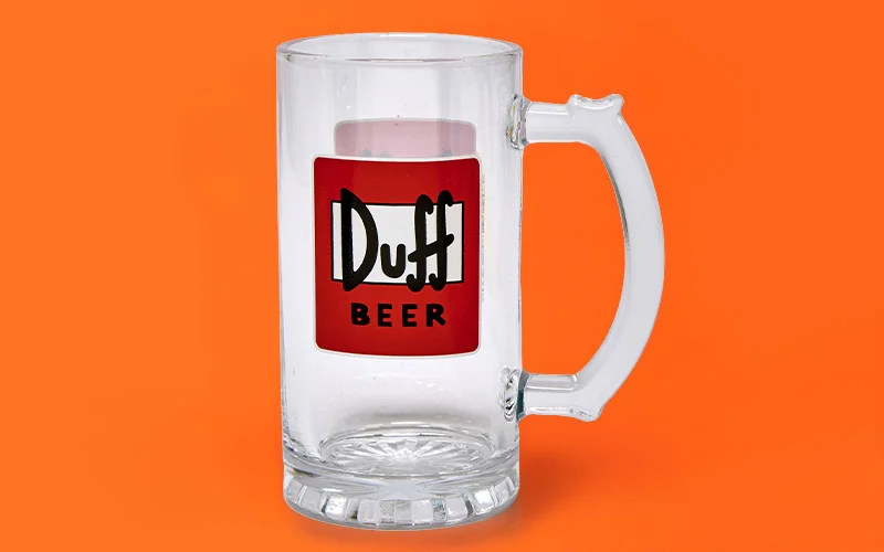 A clear glass beer tankard with a sturdy handle and thick base, featuring a red label with white and black text reading ‘Duff Beer,’ displayed against a bright orange background. A clear glass beer tankard with a sturdy handle and thick base, featuring a red label with white and black text reading ‘Duff Beer,’ displayed against a bright orange background.