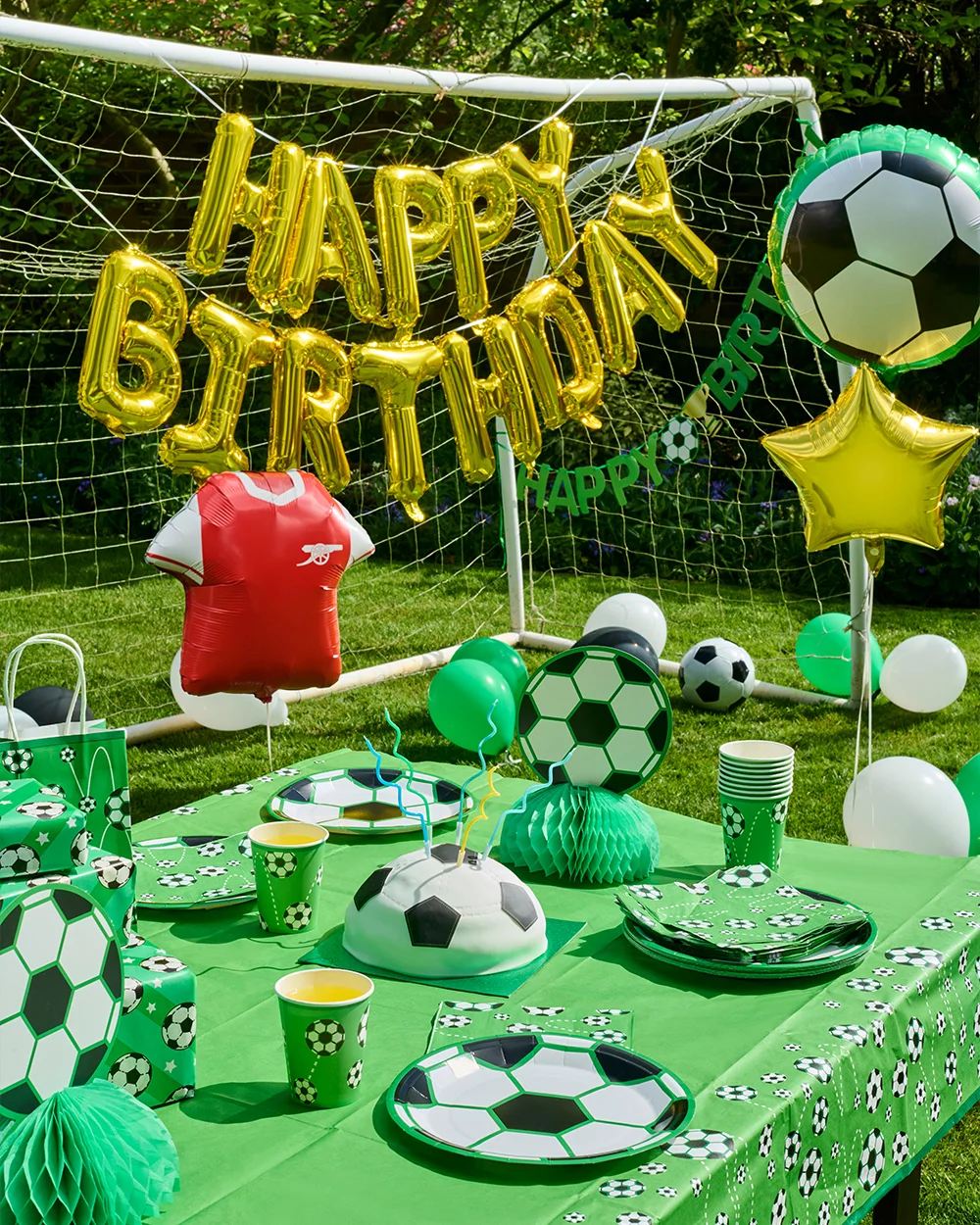 A football-themed children's party table set up outdoors on a lawn in front of a goal net, decorated with green football-print tableware including plates, cups, napkins and a matching tablecloth. The display features a football-shaped birthday cake, football honeycomb centrepieces, football-patterned gift bags and party boxes, with large gold foil ‘Happy Birthday’ balloons attached to the goal. Additional decorations include football and star-shaped foil balloons, green, white and black balloons, and a red football shirt-shaped balloon, creating a coordinated football party theme. A football-themed children's party table set up outdoors on a lawn in front of a goal net, decorated with green football-print tableware including plates, cups, napkins and a matching tablecloth. The display features a football-shaped birthday cake, football honeycomb centrepieces, football-patterned gift bags and party boxes, with large gold foil ‘Happy Birthday’ balloons attached to the goal. Additional decorations include football and star-shaped foil balloons, green, white and black balloons, and a red football shirt-shaped balloon, creating a coordinated football party theme.