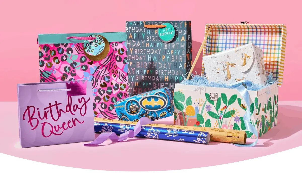 Colourful selection of birthday gift bags, boxes, and wrap displayed against a pink background.