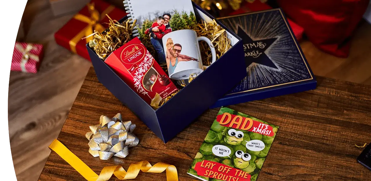 Festive holiday display with greeting cards, themed mugs labeled Mummy Claus, Daddy Claus, and Mini Claus, candles, pen, gold ribbon, and Christmas ornaments against a red background. Festive holiday display with greeting cards, themed mugs labeled Mummy Claus, Daddy Claus, and Mini Claus, candles, pen, gold ribbon, and Christmas ornaments against a red background.