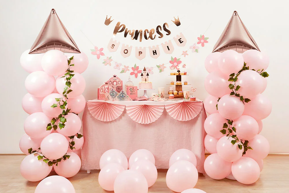 A princess-themed celebration display featuring a pink party table with fan decorations along the front, surrounded by pastel pink balloon towers with rose-gold castle-turret balloons. A personalised ‘Princess Sophie’ banner hangs above the table alongside floral garlands, with cakes, cupcakes and princess-themed party accessories arranged below. A princess-themed celebration display featuring a pink party table with fan decorations along the front, surrounded by pastel pink balloon towers with rose-gold castle-turret balloons. A personalised ‘Princess Sophie’ banner hangs above the table alongside floral garlands, with cakes, cupcakes and princess-themed party accessories arranged below.