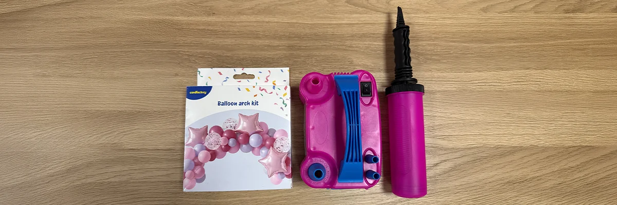 AA balloon arch kit displayed on a wooden surface, featuring the kit packaging illustrated with a pink balloon arch design, a pink electric balloon pump with blue controls, and a matching hand pump for inflating balloons. A balloon arch kit displayed on a wooden surface, featuring the kit packaging illustrated with a pink balloon arch design, a pink electric balloon pump with blue controls, and a matching hand pump for inflating balloons.