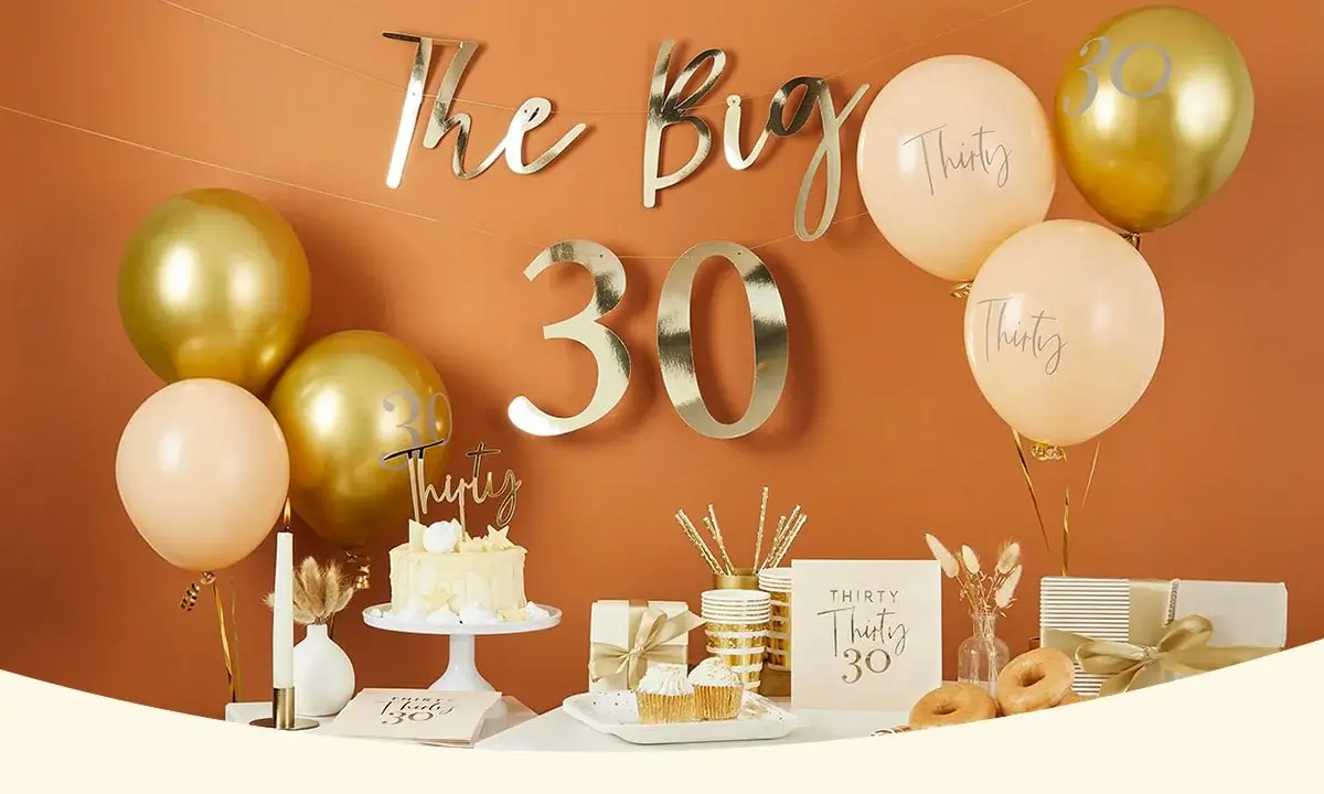 30th birthday celebration setup with gold and peach decorations, including balloons with 'Thirty', a cake, cupcakes, donuts, wrapped gifts, and a sign reading 'The Big 30' against an orange wall.