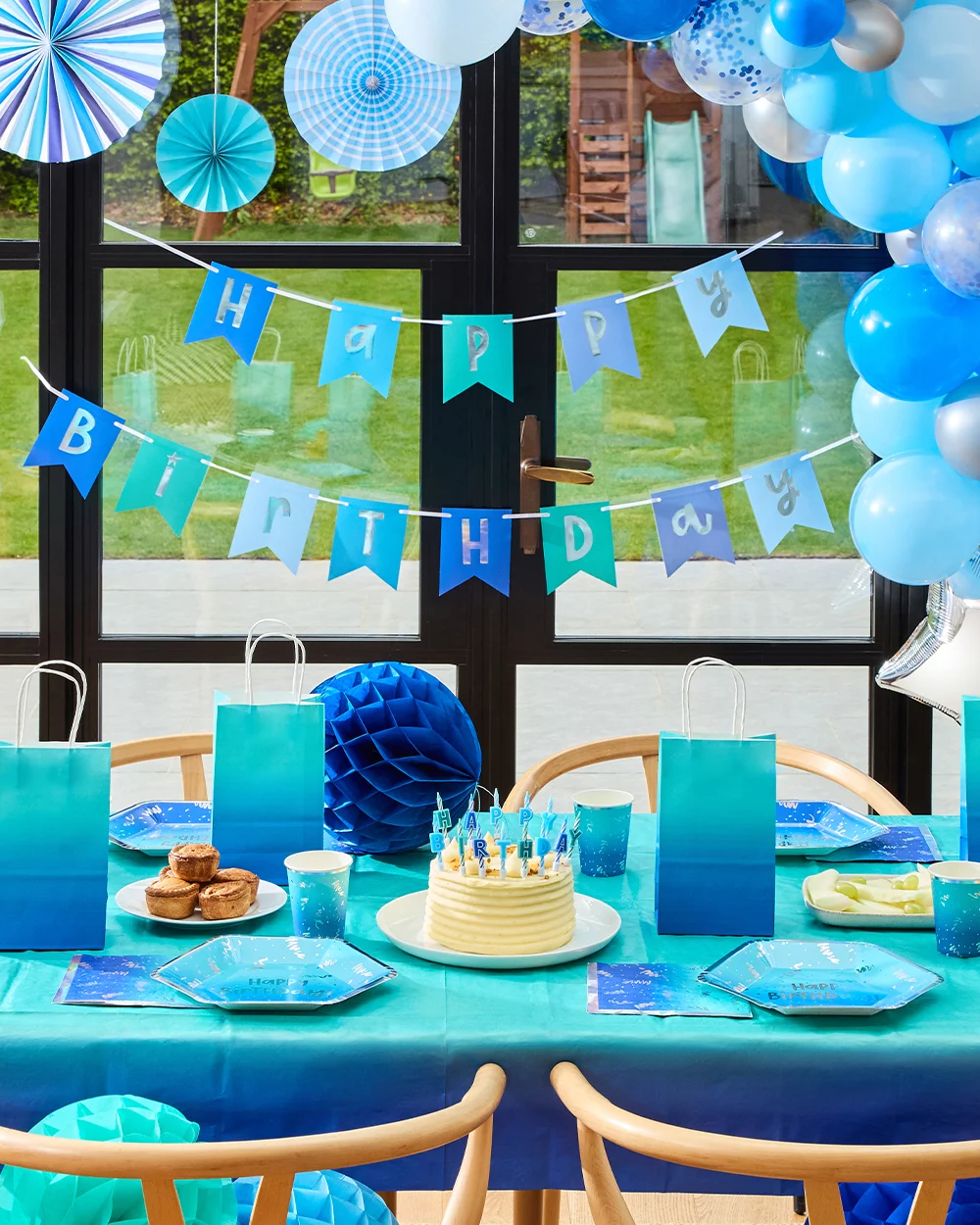 A blue-themed birthday party table set in front of glass patio doors, decorated with a blue and silver balloon garland, blue paper fan decorations and a ‘Happy Birthday’ bunting banner in shades of blue. The table is styled with blue party plates, cups and napkins, ombre blue party bags, a birthday cake topped with candles, and party food including cupcakes and fruit, creating a coordinated blue celebration theme.
