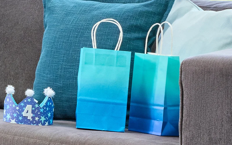A pair of blue ombré paper party bags with white handles displayed on a sofa beside blue cushions. A blue crown-shaped party hat decorated with stars, white pom-poms and a number 4 sits alongside the bags, creating a simple children's birthday party favour display. A pair of blue ombré paper party bags with white handles displayed on a sofa beside blue cushions. A blue crown-shaped party hat decorated with stars, white pom-poms and a number 4 sits alongside the bags, creating a simple children's birthday party favour display.