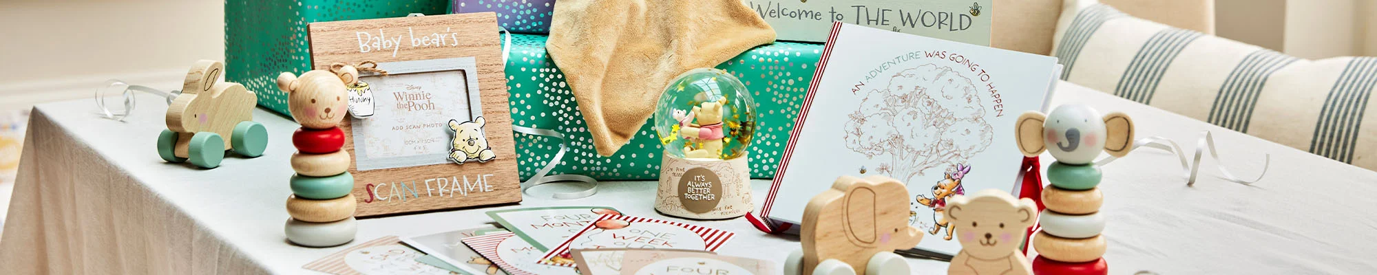 A literary-themed baby gift display featuring Winnie the Pooh keepsakes, baby books, wooden stacking toys and wrapped presents arranged on a table. The display includes a Winnie the Pooh scan frame, a ‘Welcome to the World’ photo frame, a snow globe, greeting cards, and baby-themed gifts set against a bright indoor backdrop with white balloons.” A literary-themed baby gift display featuring Winnie the Pooh keepsakes, baby books, wooden stacking toys and wrapped presents arranged on a table. The display includes a Winnie the Pooh scan frame, a ‘Welcome to the World’ photo frame, a snow globe, greeting cards, and baby-themed gifts set against a bright indoor backdrop with white balloons.”