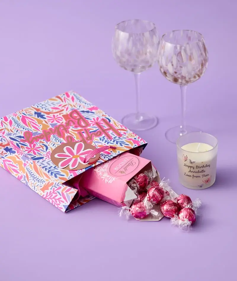 Birthday gift set with a floral gift bag, chocolates, personalised candle, and decorative wine glasses on a lilac background.