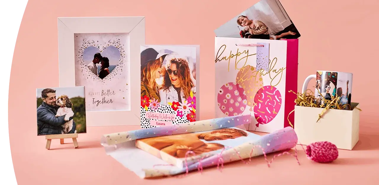 A display of personalised photo gifts on a peach background, including a photo frame, greeting cards, a birthday gift bag, a photo mug, and wrapped photo prints.