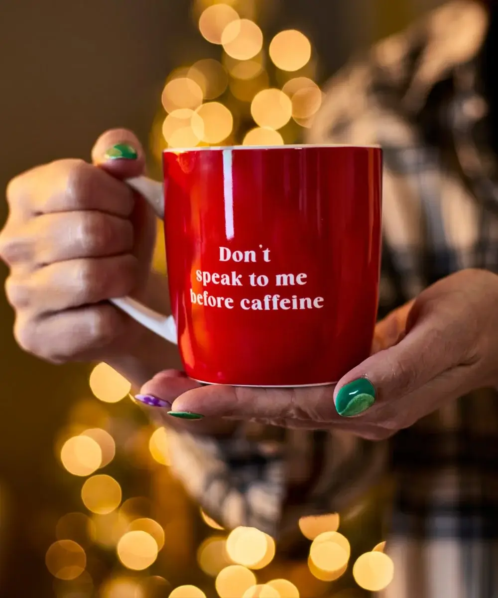 Person holding a red coffee mug with the text Don’t speak to me before caffeine, with festive lights blurred in the background.