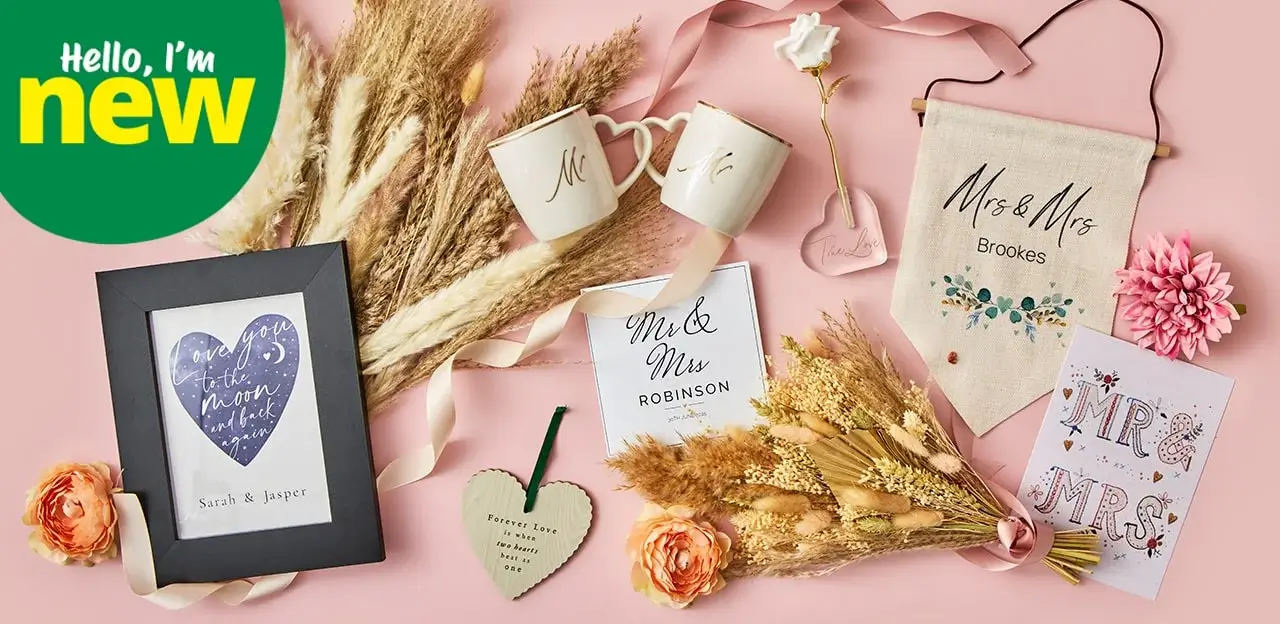 The image shows a collection of wedding-themed items arranged on a pink surface. There are two white mugs with 