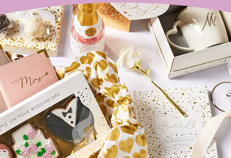 'A wedding-themed flat lay featuring various items including a pink notebook with the word 'Mrs.' in gold script, a bottle of champagne labeled 'Cheers', heart-shaped cookies, a white mug with an 'M' on it, and a journal titled 'Our Bucket List: An Adventure Journal for Couples'. The items are surrounded by gold and white wrapping paper with heart patterns.'