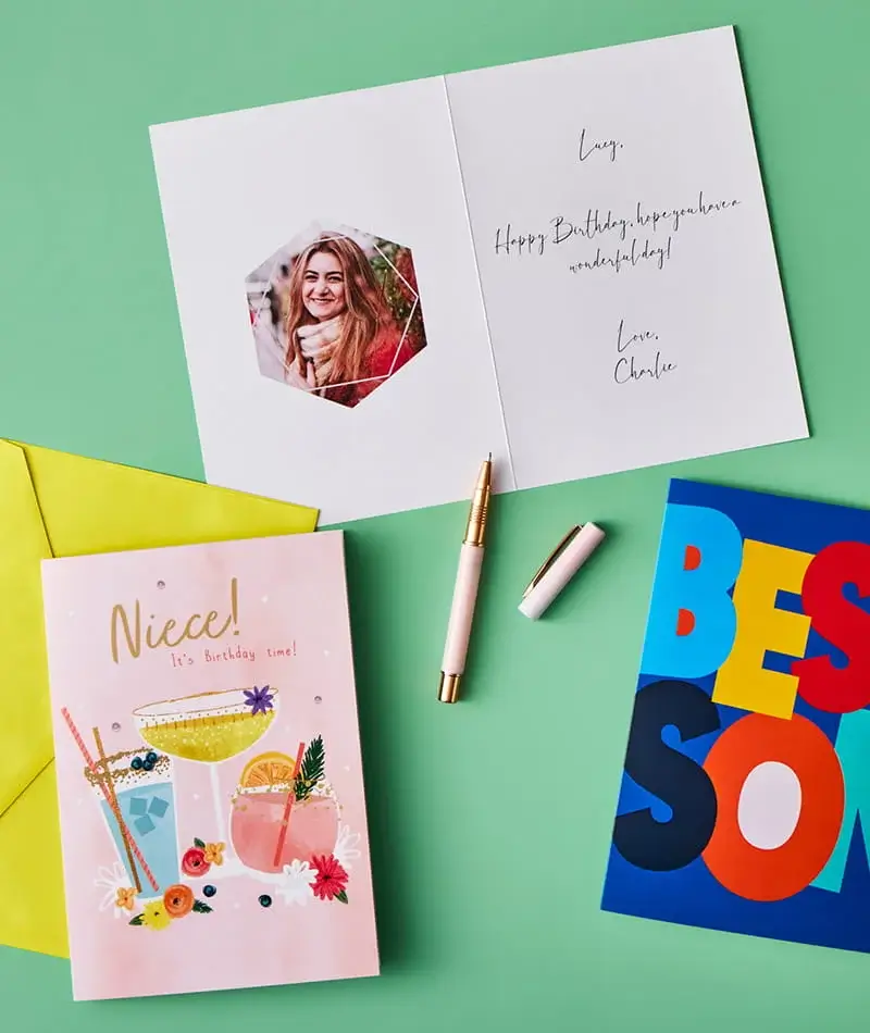 Open birthday card with a photo insert and handwritten message, surrounded by colourful cards, envelopes, and pens on a green background