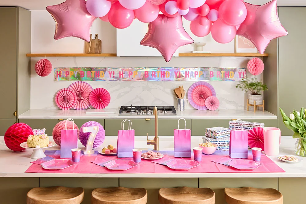 A pastel pink birthday party setup in a modern kitchen featuring a pink balloon garland with star-shaped foil balloons, a holographic ‘Happy Birthday’ banner, paper fan decorations and matching pink partyware. The table is arranged with ombré party bags, plates, cups, wrapped presents, snacks and a birthday cake beside a light-up number 7 decoration. A pastel pink birthday party setup in a modern kitchen featuring a pink balloon garland with star-shaped foil balloons, a holographic ‘Happy Birthday’ banner, paper fan decorations and matching pink partyware. The table is arranged with ombré party bags, plates, cups, wrapped presents, snacks and a birthday cake beside a light-up number 7 decoration.