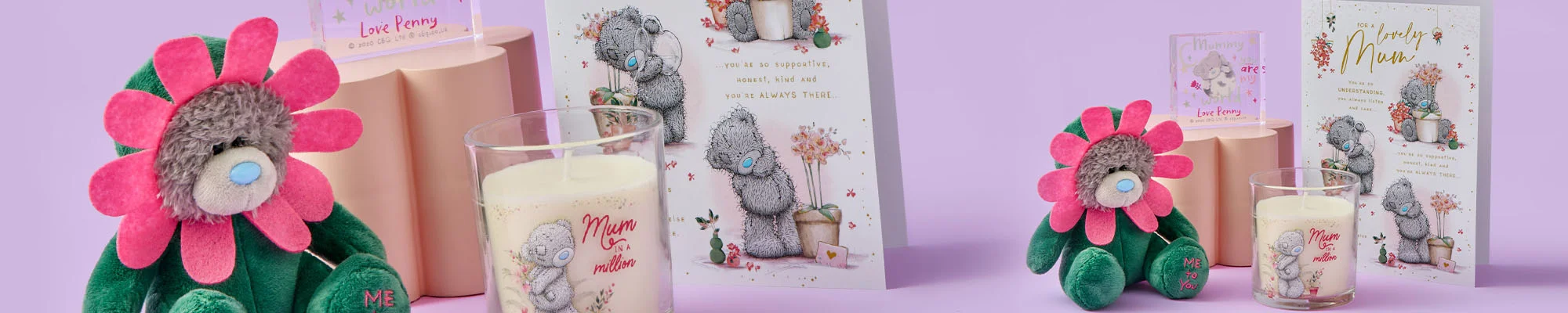 A wide display of Tatty Teddy Mother’s Day gifts, including a flower‑costumed plush toy, a scented candle, a decorative acrylic block, and a greeting card, arranged on a pastel purple background.