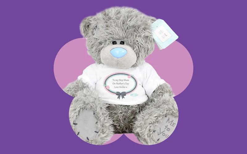 A grey Tatty Teddy bear wearing a white T‑shirt printed with a Mother’s Day message, displayed against a purple background with a pink flower shape.