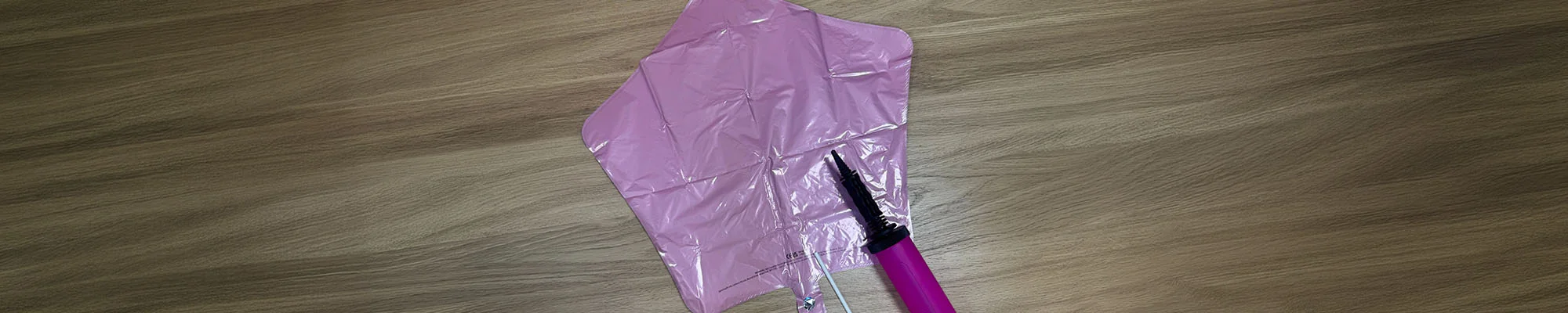 A pink star-shaped foil balloon laid flat on a wooden surface beside a hand balloon pump, showing how the balloon is inflated. The balloon is attached to a white balloon stick, demonstrating the components included in a balloon decoration kit. A pink star-shaped foil balloon laid flat on a wooden surface beside a hand balloon pump, showing how the balloon is inflated. The balloon is attached to a white balloon stick, demonstrating the components included in a balloon decoration kit.