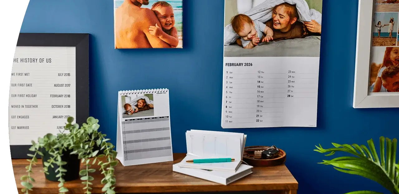 Personalised photo calendars and keepsake prints displayed on a desk with plants, notebooks, and framed family photographs against a blue wall. TPersonalised photo calendars and keepsake prints displayed on a desk with plants, notebooks, and framed family photographs against a blue wall.