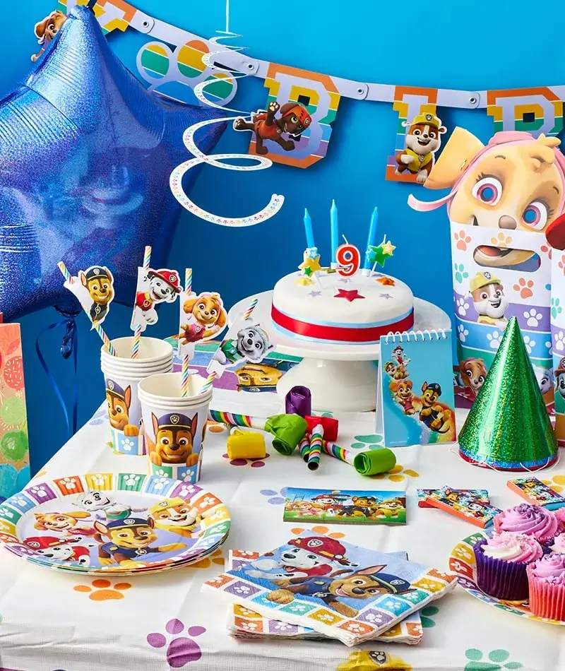 A colorful birthday party setup featuring Paw Patrol-themed decorations. The table has a white tablecloth with paw prints, Paw Patrol plates, cups, and napkins. A cake with blue candles and a number 9 topper is surrounded by party favors like blowouts and hats. A large blue star balloon and a 'HAPPY BIRTHDAY' banner hang in the background. A colorful birthday party setup featuring Paw Patrol-themed decorations. The table has a white tablecloth with paw prints, Paw Patrol plates, cups, and napkins. A cake with blue candles and a number 9 topper is surrounded by party favors like blowouts and hats. A large blue star balloon and a 'HAPPY BIRTHDAY' banner hang in the background.