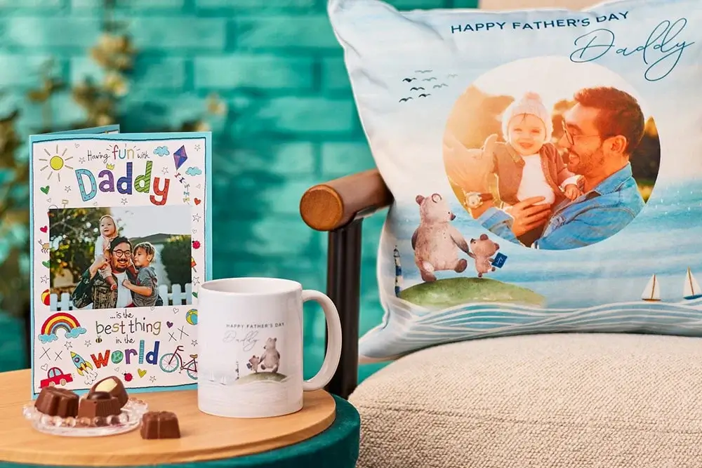 The image shows a Father's Day setup with a personalised card, mug, and pillow. The card has colourful drawings and text that reads 'Having fun with Daddy is the best thing in the world.' The mug features a picture of a father and child with the text 'Happy Father's Day.' The pillow has an image of a father holding his child, with the text 'HAPPY FATHER'S DAY Daddy' at the top. There are also chocolates on the table next to these items. The image shows a Father's Day setup with a personalised card, mug, and pillow. The card has colourful drawings and text that reads 'Having fun with Daddy is the best thing in the world.' The mug features a picture of a father and child with the text 'Happy Father's Day.' The pillow has an image of a father holding his child, with the text 'HAPPY FATHER'S DAY Daddy' at the top. There are also chocolates on the table next to these items.