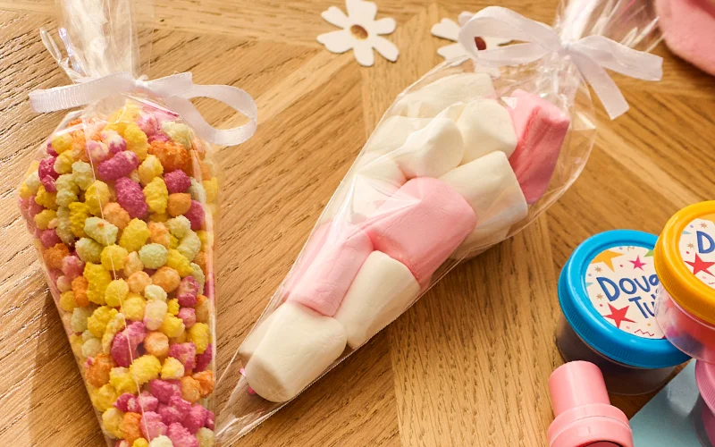 Two clear sweet cones tied with white ribbon, one filled with colourful sugar-coated popcorn and the other containing pink and white marshmallows. The treats are displayed on a wooden tabletop beside party bag filler items and daisy-shaped decorations. Two clear sweet cones tied with white ribbon, one filled with colourful sugar-coated popcorn and the other containing pink and white marshmallows. The treats are displayed on a wooden tabletop beside party bag filler items and daisy-shaped decorations.