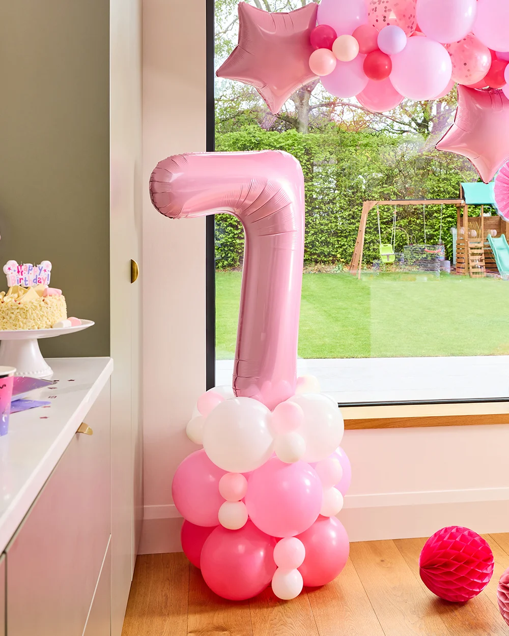 A princess-themed balloon centrepiece featuring a large pastel pink number 7 foil balloon mounted on a base of pink, white and fuchsia balloons. The display is positioned beside a party table with a birthday cake, while pink star-shaped foil balloons and a balloon garland decorate the room in the background. A princess-themed balloon centrepiece featuring a large pastel pink number 7 foil balloon mounted on a base of pink, white and fuchsia balloons. The display is positioned beside a party table with a birthday cake, while pink star-shaped foil balloons and a balloon garland decorate the room in the background.