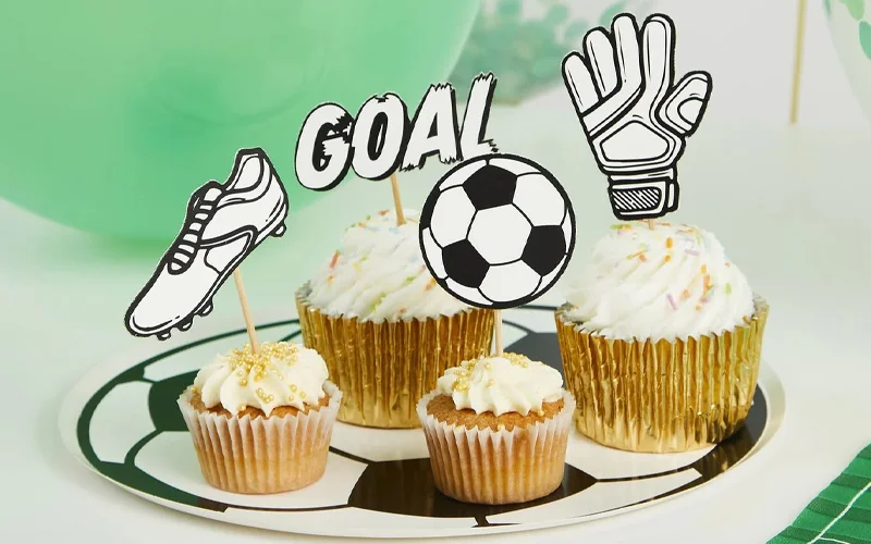 A selection of cupcakes displayed on a football-patterned plate, decorated with football-themed toppers including a football boot, goalkeeper glove, football and the word ‘GOAL.’ A selection of cupcakes displayed on a football-patterned plate, decorated with football-themed toppers including a football boot, goalkeeper glove, football and the word ‘GOAL.’