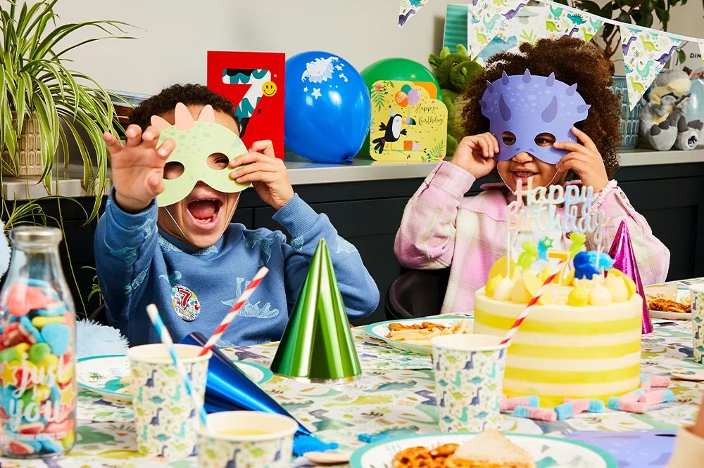 A dinosaur-themed birthday party scene featuring children wearing dinosaur masks seated at a decorated party table. The display includes dinosaur-print tableware, party hats, balloons, a birthday cake with a number 7 candle, dinosaur bunting and themed birthday decorations.