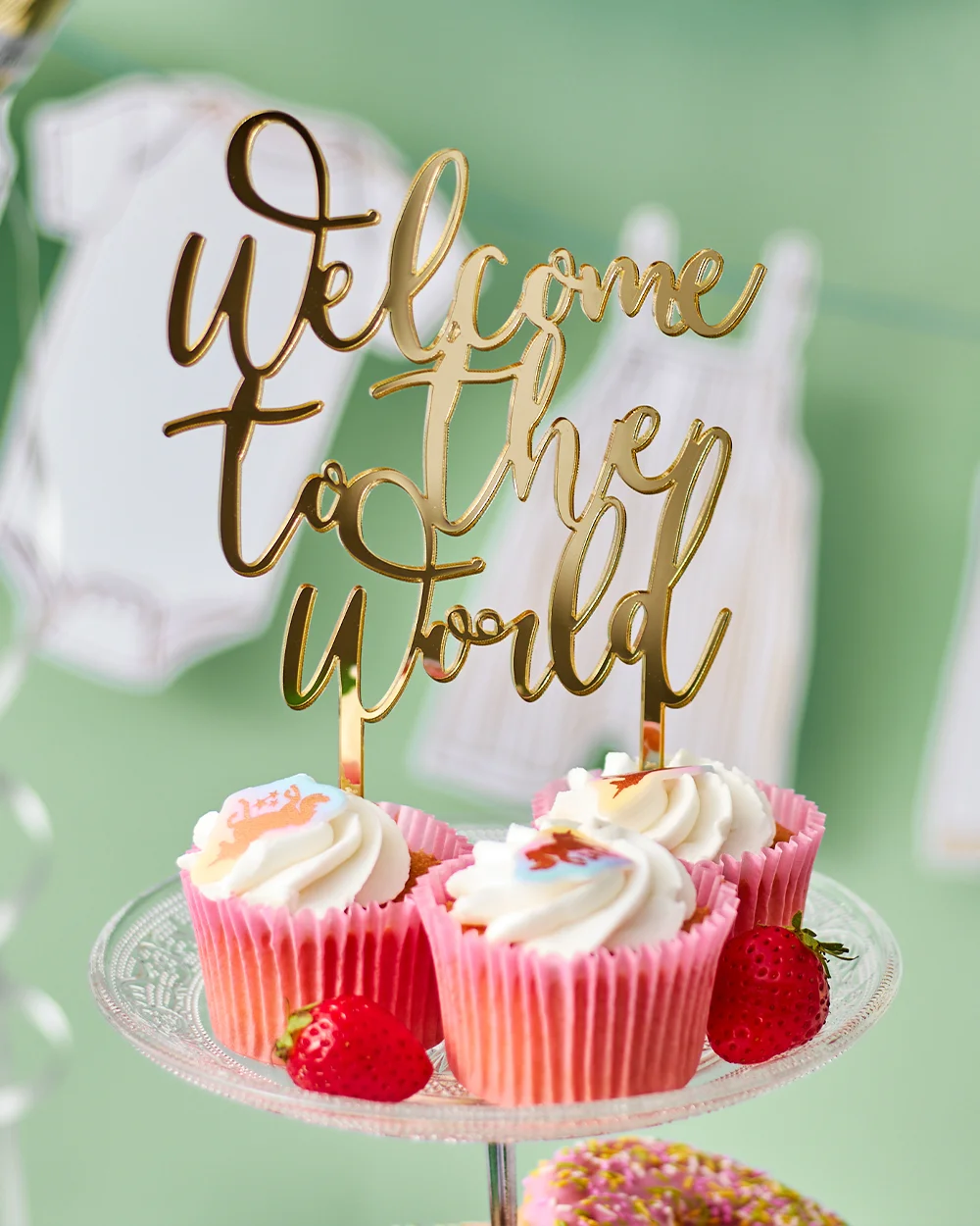 A baby shower cupcake stand featuring pink cupcakes topped with white icing and baby-themed decorations, displayed around a gold script sign reading ‘Welcome to the World.’ Fresh strawberries and pink-sprinkled doughnuts are arranged on the stand, with baby clothes bunting and a sage green backdrop completing the baby shower dessert display.