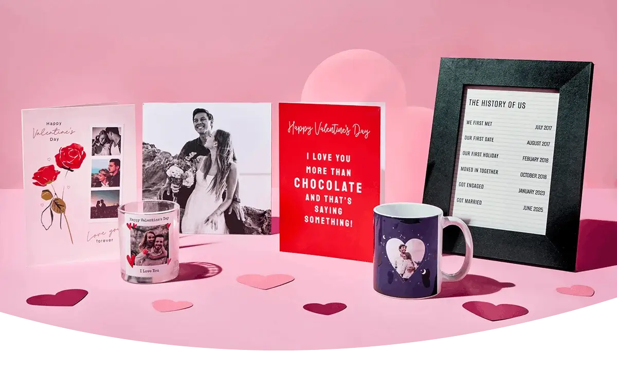 A display of Valentine’s gifts on a pink surface, including greeting cards, two photo mugs, heart‑shaped confetti, and a framed timeline titled The History of Us.