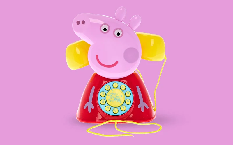 A Peppa Pig character‑shaped toy telephone with a rotary‑style dial and yellow handset, set against a pink background.