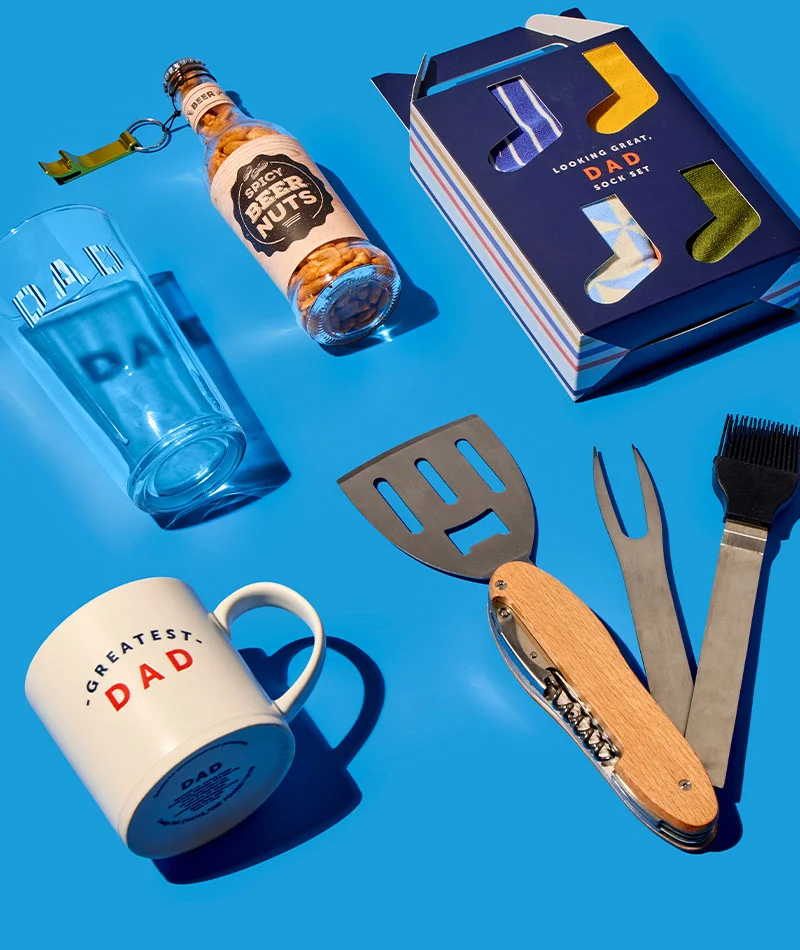 A bright blue flat‑lay display of Father’s Day gifts, including a clear pint glass printed ‘DAD,’ a bottle of ‘Spicy Beer Nuts’ with a keyring bottle opener attached, a navy ‘Looking Great, Dad’ sock set box with colourful socks visible, a cream mug reading ‘Greatest Dad,’ and a set of barbecue tools with wooden handles laid out beside them. A bright blue flat‑lay display of Father’s Day gifts, including a clear pint glass printed ‘DAD,’ a bottle of ‘Spicy Beer Nuts’ with a keyring bottle opener attached, a navy ‘Looking Great, Dad’ sock set box with colourful socks visible, a cream mug reading ‘Greatest Dad,’ and a set of barbecue tools with wooden handles laid out beside them.